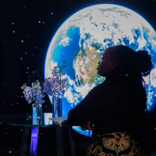 Naom Sang wearing a black, long sleeved top and yellow and black  pants, leans on a glass table with floral arrangements, thoughtfully facing a large digital display of Earth
